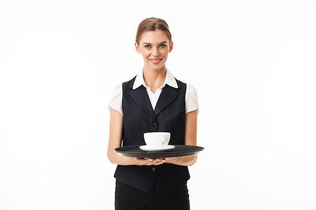 Giovane bella cameriera in uniforme tenendo il vassoio con una tazza di caffè mentre felicemente