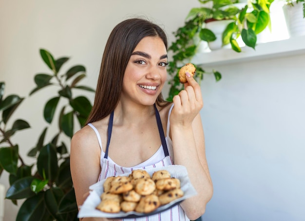 Giovane bella casalinga che tiene i biscotti appena sfornati su un vassoio in cucina