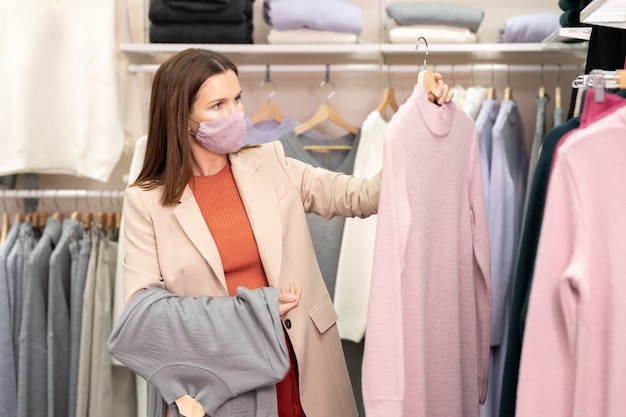 Giovane bella cliente bruna in maschera e abbigliamento casual intelligente in piedi accanto al rack con un nuovo assortimento di vestiti caldi e scegliendo un abito elegante