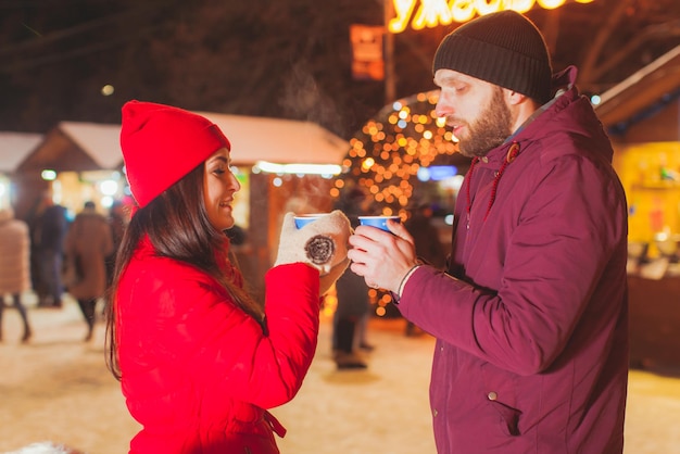 Giovane bella coppia in fase di riscaldamento al mercatino di Natale, tenendo le tazze con tè caldo, in piedi di fronte e guardandosi.