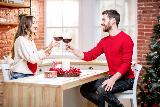 Giovane bella coppia vestita di maglioni che festeggiano il nuovo anno bevendo vino nell'appartamento con l'albero di Natale