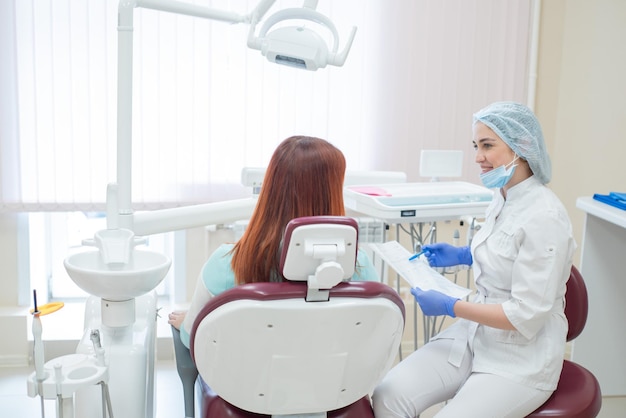 Giovane bella dentista femminile che parla con un paziente in ufficio Una donna dai capelli rossi siede su una sedia con un ortodontista e ascolta gli appuntamenti Servizi odontoiatrici Vista dal retro