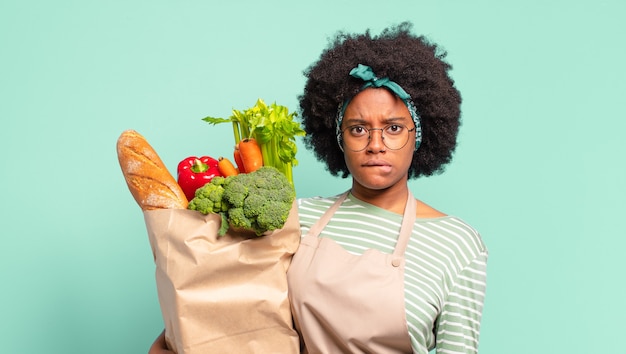 Giovane bella donna afro che sembra perplessa e confusa, mordendosi il labbro con un gesto nervoso, non conoscendo la risposta al problema e tenendo in mano un sacchetto di verdure