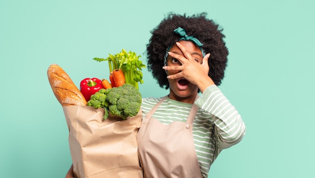 Giovane bella donna afro che si sente felice, sorpresa e orgogliosa, indicando se stessa con uno sguardo eccitato e stupito e tenendo in mano un sacchetto di verdure