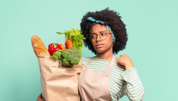 Giovane bella donna afro che sorride con orgoglio e sicurezza facendo posare trionfante il numero uno, sentendosi come un leader e tenendo in mano un sacchetto di verdure