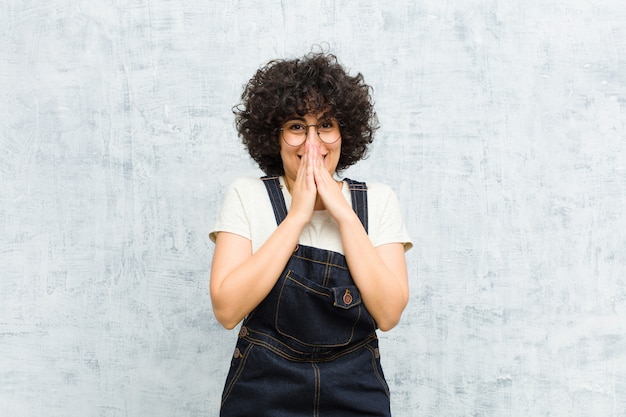 Giovane bella donna afro felice ed eccitata, sorpresa e stupita che copre la bocca con le mani, ridacchiando con un'espressione carina