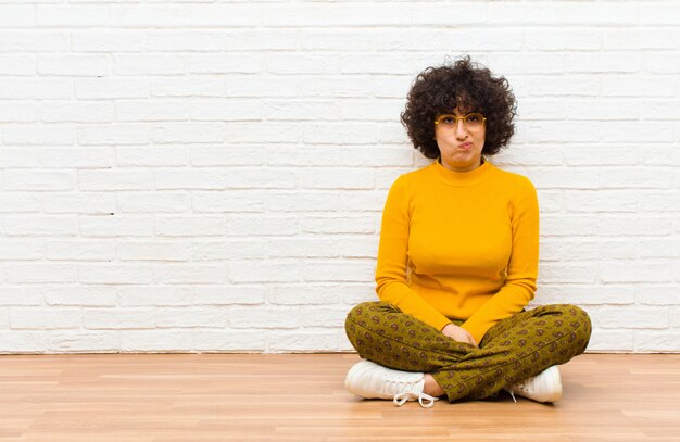 Giovane bella donna afro sentirsi confusa e dubbiosa, chiedendosi o cercando di scegliere o prendere una decisione seduta sul pavimento