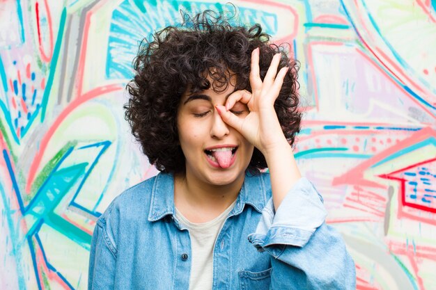 Giovane bella donna afro sorridente felicemente con la faccia buffa, scherzando e guardando attraverso lo spioncino, spiando segreti contro il muro dei graffiti