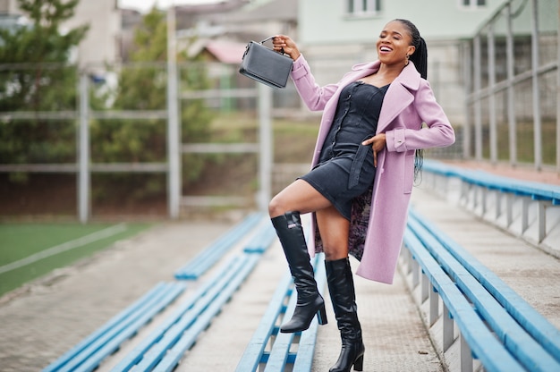 Giovane bella donna afroamericana alla moda in strada presso le gradinate dello stadio, indossando il cappotto dell'attrezzatura di moda, con la borsa.