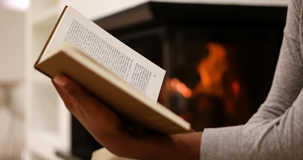 Giovane bella donna afroamericana seduta davanti al caminetto a casa in una fredda giornata autunnale e libro di lettura