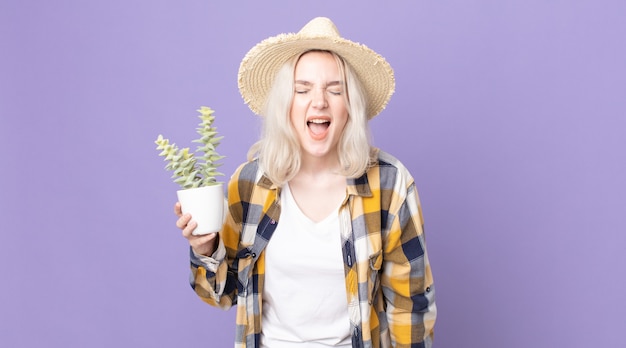 Giovane bella donna albina che grida in modo aggressivo, sembra molto arrabbiata e tiene in mano un cactus di pianta d'appartamento