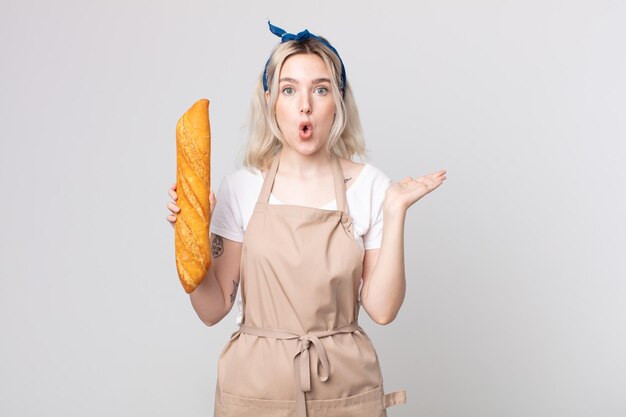 giovane bella donna albina che sembra sorpresa e scioccata, con la mascella caduta che tiene un oggetto con una baguette di pane