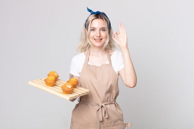 Giovane bella donna albina che si sente felice, mostra approvazione con un gesto ok con un vassoio di muffin