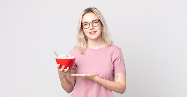 Giovane bella donna albina che sorride allegramente sentendosi felice e mostrando un concetto con una ciotola per la colazione