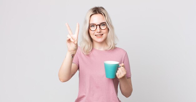 giovane bella donna albina che sorride e sembra felice, gesticolando vittoria o pace con una tazza di caffè