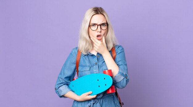 Giovane bella donna albina con la bocca e gli occhi spalancati e la mano sul mento che tiene in mano uno skateboard