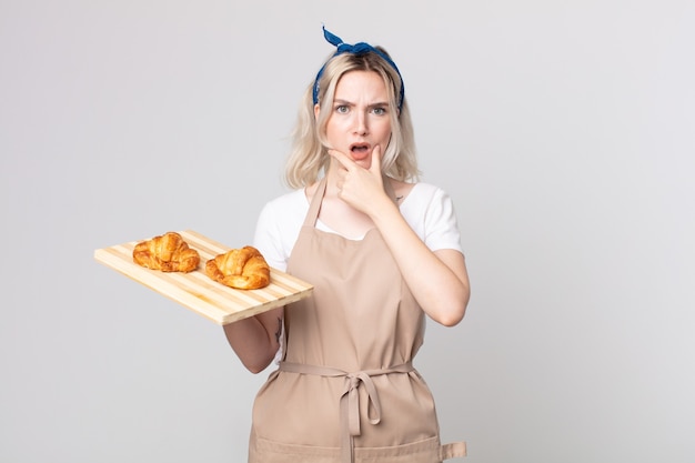 Giovane bella donna albina con la bocca e gli occhi spalancati e la mano sul mento con un vassoio di croissant