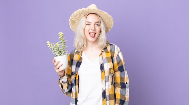 Giovane bella donna albina con un atteggiamento allegro e ribelle, che scherza e tira fuori la lingua e tiene in mano un cactus di pianta d'appartamento