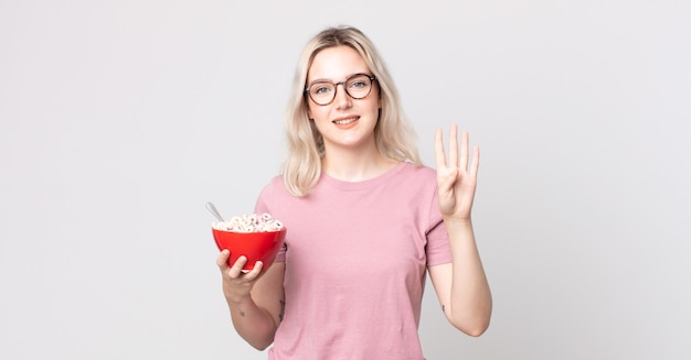 Giovane bella donna albina sorridente e dall'aspetto amichevole, che mostra il numero quattro con una ciotola per la colazione