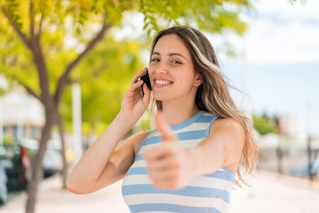 Giovane bella donna all'aperto che tiene una conversazione con il cellulare mentre fa il pollice in alto