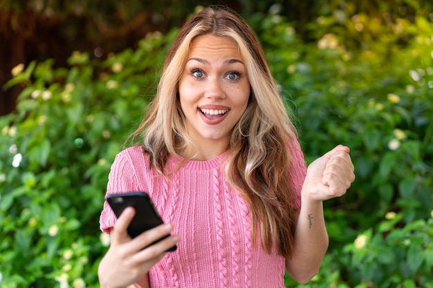 Giovane bella donna all'aperto usando il telefono cellulare e facendo il gesto della vittoria