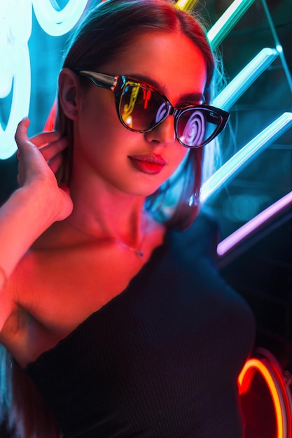 Giovane bella donna alla moda con occhiali da sole eleganti in un abito nero su sfondo scuro con luci al neon colorate e luminose Bellezza urbana della ragazza