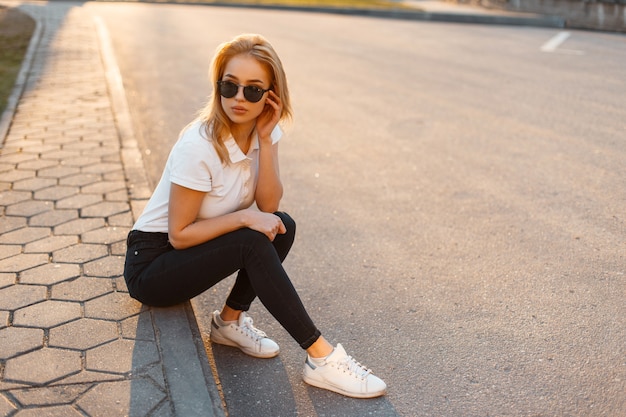 Giovane bella donna alla moda hipster con occhiali da sole in una polo bianca e jeans neri con scarpe da ginnastica che si siedono sulla strada al tramonto. Ragazza alla moda in abiti estivi