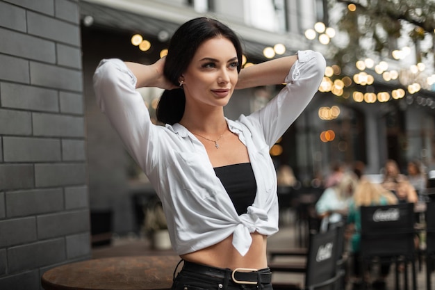 Giovane bella donna alla moda in abiti casual con camicia bianca si trova in città e fissa i capelli su uno sfondo urbano con bokeh leggero