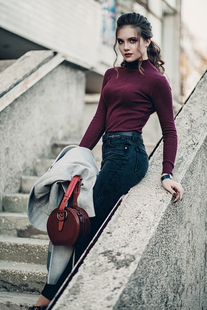 Giovane bella donna alla moda in jeans neri e con una borsa rosso scuro. Modello professionale.