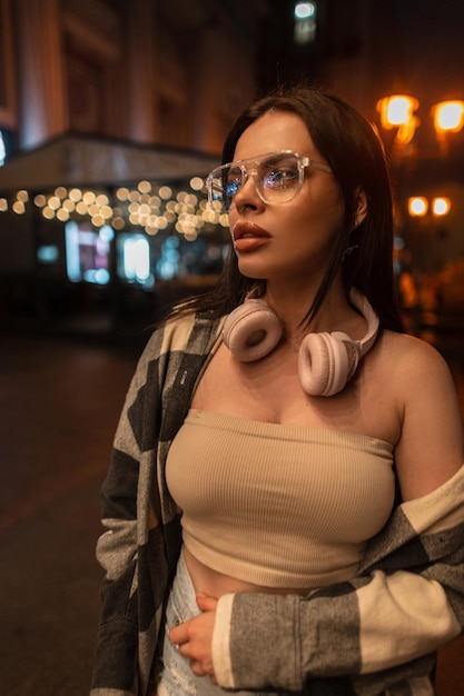 Giovane bella donna alla moda urbana con occhiali eleganti e cuffie rosa in abiti casual alla moda con top e camicia a quadri che cammina nella città notturna con luci bokeh