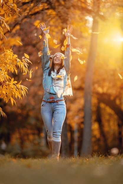 Giovane bella donna allegra che va da sola nel parco e getta le foglie in autunno soleggiato dorato.