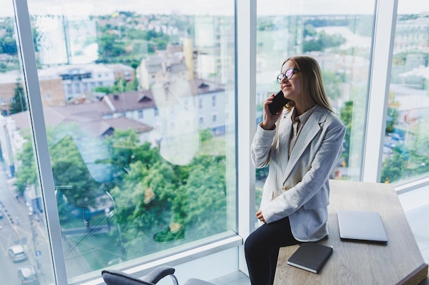 Giovane bella donna allegra con gli occhiali che parla su un telefono cellulare e utilizza un computer portatile con un sorriso mentre è seduto al suo posto di lavoro Lavoro in ufficio