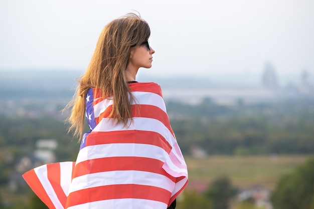 Giovane bella donna americana con i capelli lunghi che tiene sventolando la bandiera USA del vento