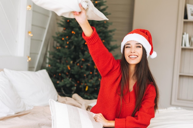 Giovane bella donna asiatica con capelli lunghi scuri in un accogliente maglione rosso lavorato a maglia e cappello da Babbo Natale divertendosi nella stanza con l'albero di Natale a casa