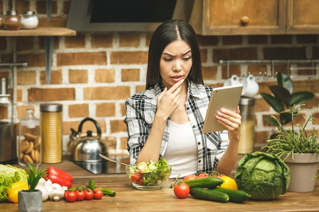 Giovane bella donna asiatica con la compressa sulla cucina che trova le ricette e sorridere. Avvicinamento.