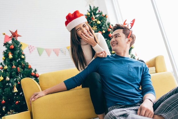 Giovane bella donna asiatica felice che indossa il cappello di Babbo Natale sorprende e ride nella parte posteriore del suo ragazzo a casa con un albero di Natale sullo sfondo Immagine con spazio di copia