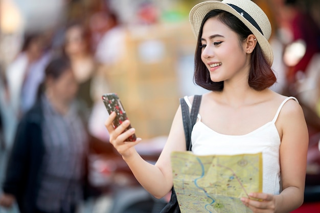 Giovane bella donna asiatica guardando lo schermo dello smartphone e tenendo la mappa in mano