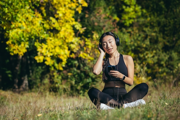 Giovane bella donna asiatica seduta nella posizione del loto nel parco, rilassarsi dopo il fitness e correre, ascoltare musica con le cuffie e utilizzare l'app mobile sul telefono per il fitness