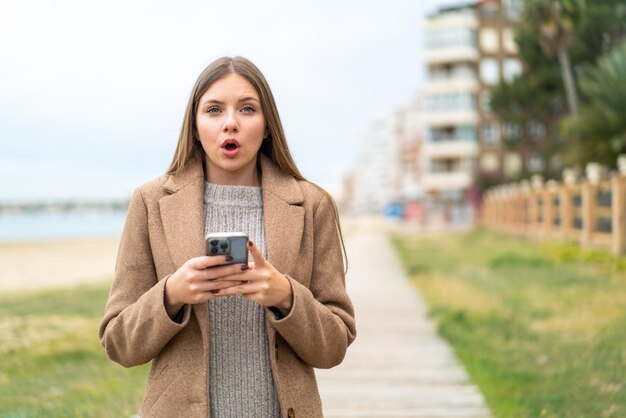 Giovane bella donna bionda che guarda la telecamera mentre utilizza il cellulare con espressione sorpresa