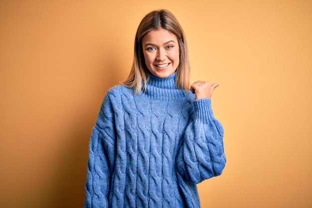 Giovane bella donna bionda che indossa un maglione a collo alto su sfondo giallo isolato sorridente con la faccia felice guardando e indicando il lato con il pollice in su