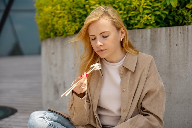 Giovane bella donna bionda che mangia sushi all'aperto sulla terrazza in legno vicino a un edificio moderno in città Cibo gustoso da portare La ragazza ha la pausa pranzo trascorrendo del tempo fuori e mangiando cibo asiatico Vita di città