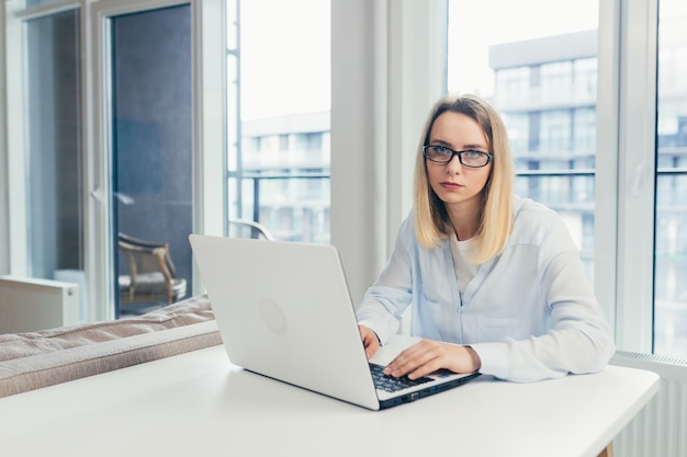 Giovane bella donna bionda che scrive su un computer portatile in ufficio stanca guardando la telecamera