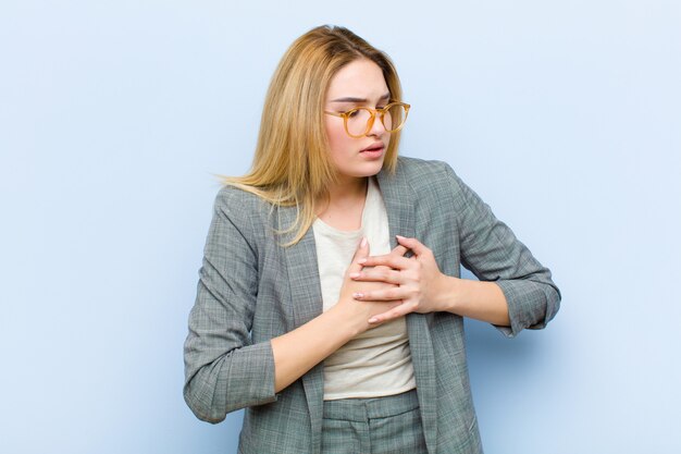 Giovane bella donna bionda che sembra triste, ferita e con il cuore spezzato, tenendo entrambe le mani vicino al cuore, piangendo e sentendosi depressa sulla parete piatta