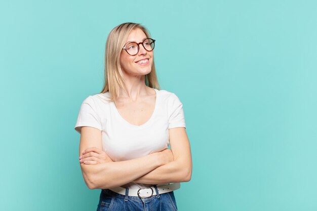 Giovane bella donna bionda che si sente felice, orgogliosa e piena di speranza, chiedendosi o pensando, alzando lo sguardo per copiare lo spazio con le braccia incrociate