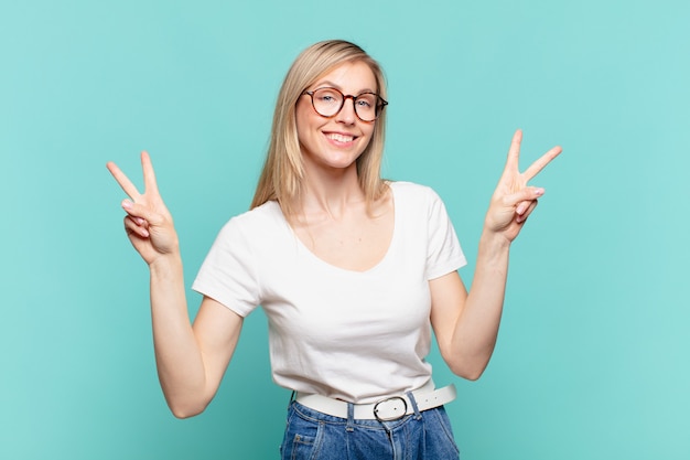 Giovane bella donna bionda che sorride e sembra felice, amichevole e soddisfatta, gesticolando vittoria o pace con entrambe le mani