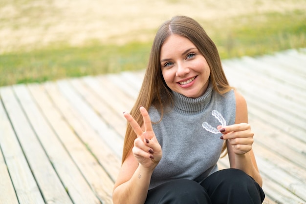 Giovane bella donna bionda che tiene le parentesi graffe invisibili all'aperto sorridendo e mostrando il segno della vittoria