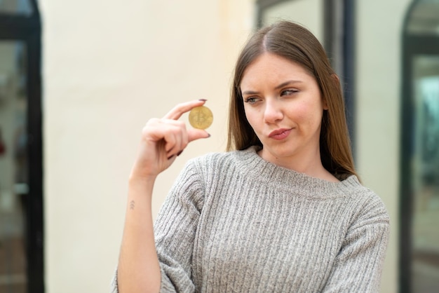 Giovane bella donna bionda che tiene un Bitcoin all'aperto con espressione triste