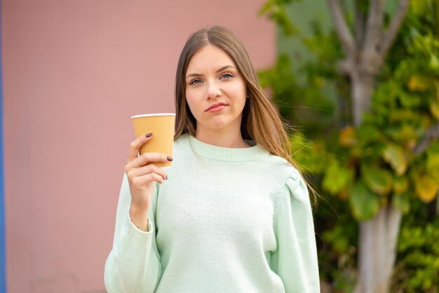 Giovane bella donna bionda che tiene un caffè da asporto all'aperto con espressione triste