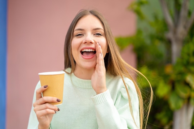 Giovane bella donna bionda che tiene un caffè da asporto all'aperto gridando con la bocca spalancata