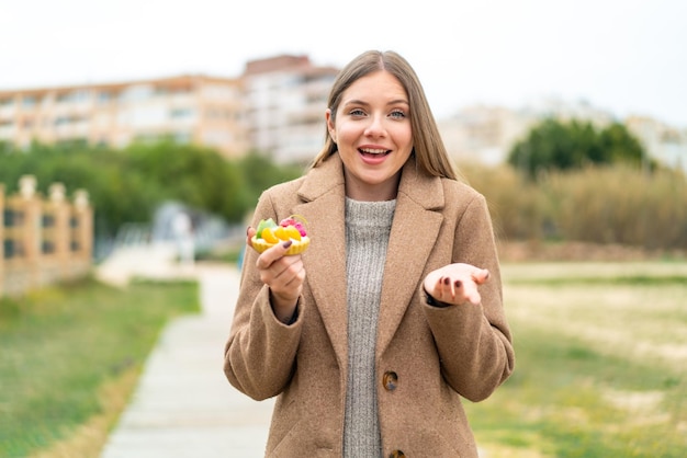 Giovane bella donna bionda che tiene un tortino all'aperto con un'espressione facciale scioccata